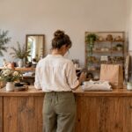 A boutique owner seen from behind stands at a wooden counter holding a smartphone, with a folded linen shirt, a paper bag, a notebook and a mug beside them